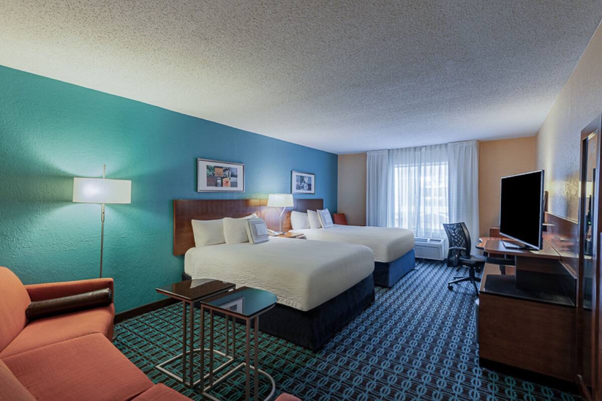 A hotel room with two beds, a desk, a chair, a TV, and a lamp.