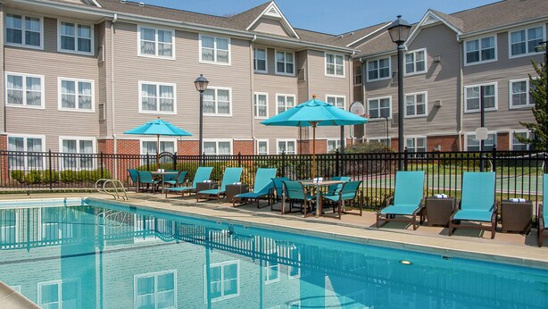 A swimming pool with lounge chairs and umbrellas, surrounded by a fence and a row of apartment buildings in the background.