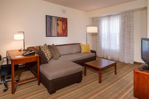 A hotel room with a sofa, a desk with a lamp, a television, and a window with curtains.