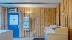 Een kamer met een houten wand, een blauwe deur, een zeep- en bleekmiddeldispenser aan de muur, en een klein tafeltje met stoelen.