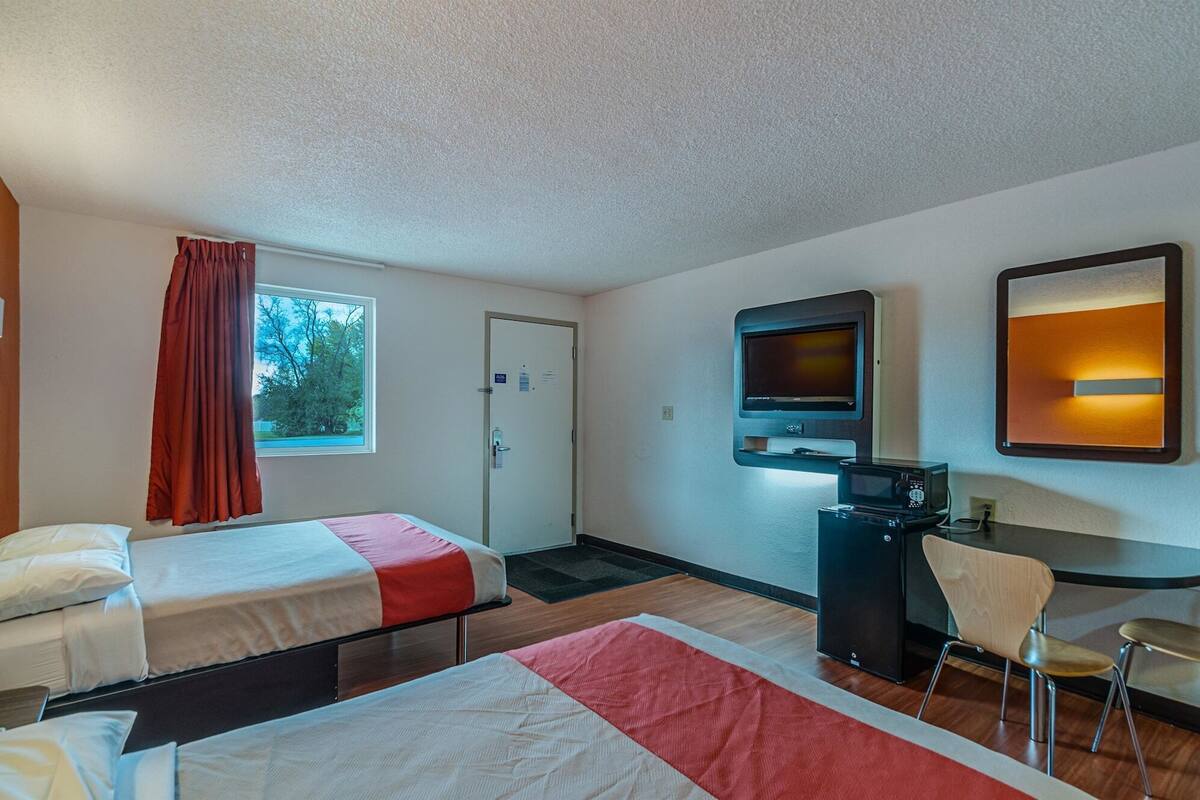 A hotel room with two beds, a TV, a desk with a chair, and a door.