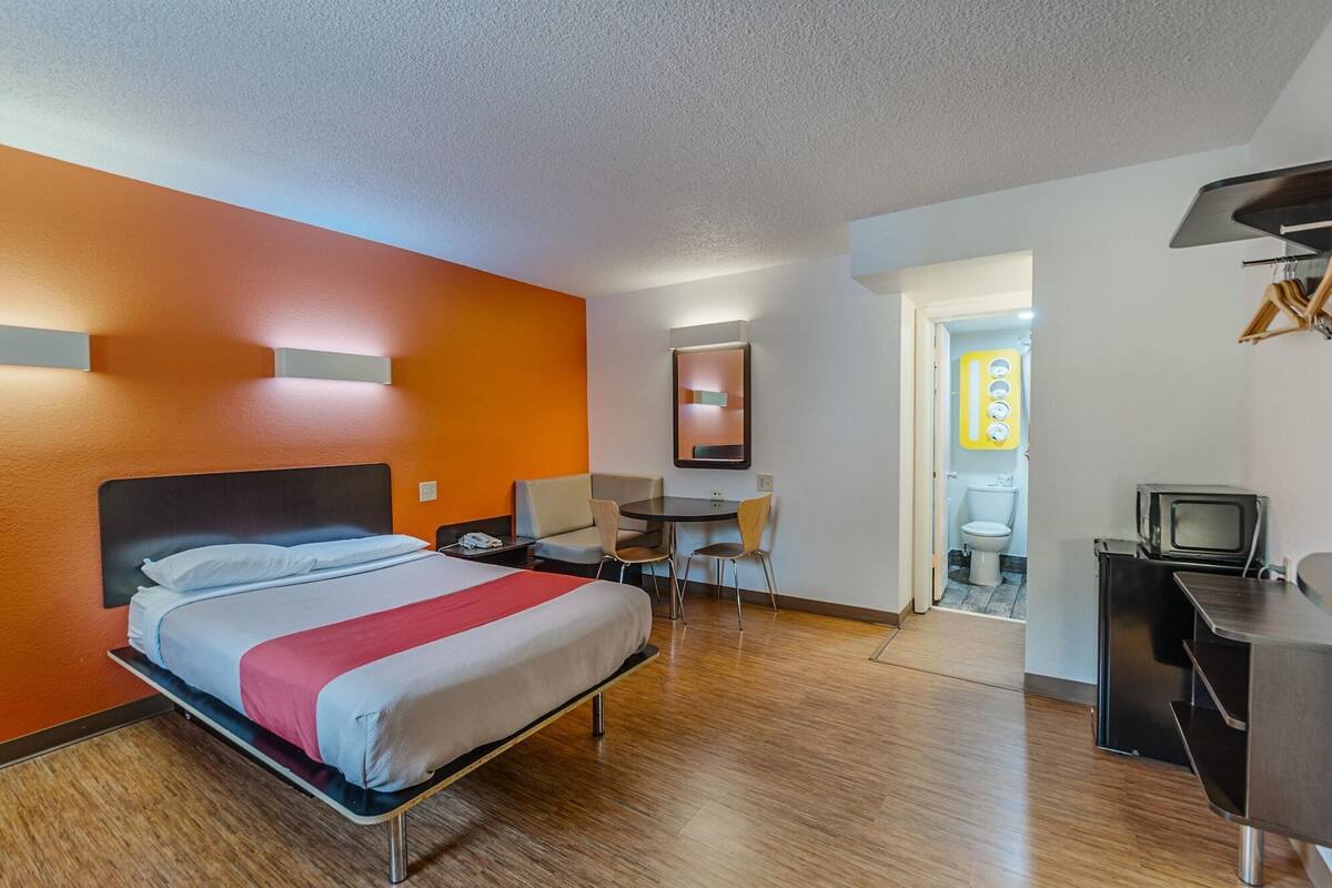 A hotel room with a bed, a desk, a chair, a small table, a television, and a bathroom visible through an open door.
