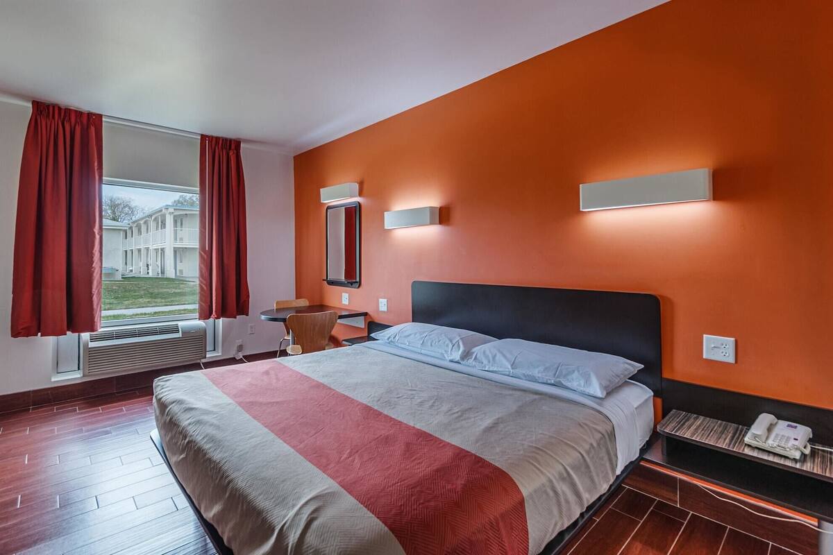 A hotel room with a large bed, a desk with a chair, a small table, a telephone, and a window with curtains.