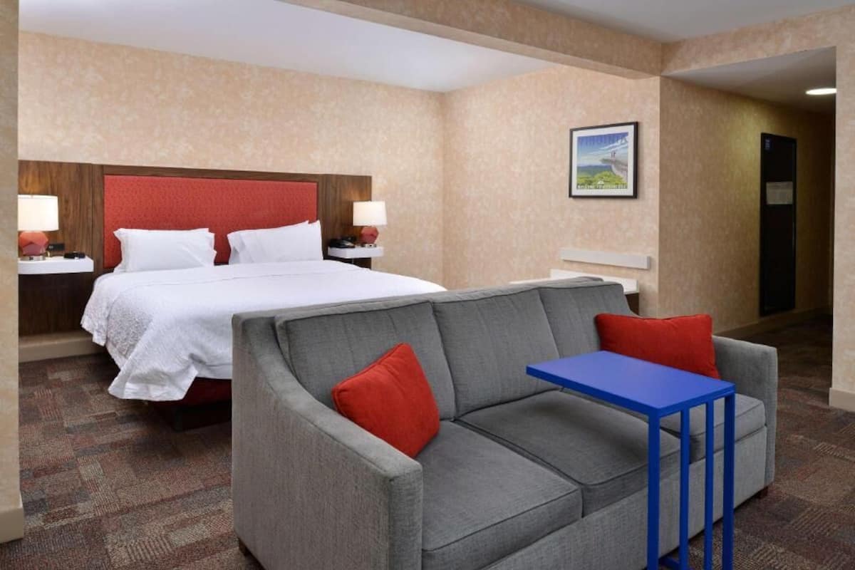 A hotel room with a bed, a grey sofa, a small blue table, and a nightstand with a lamp.