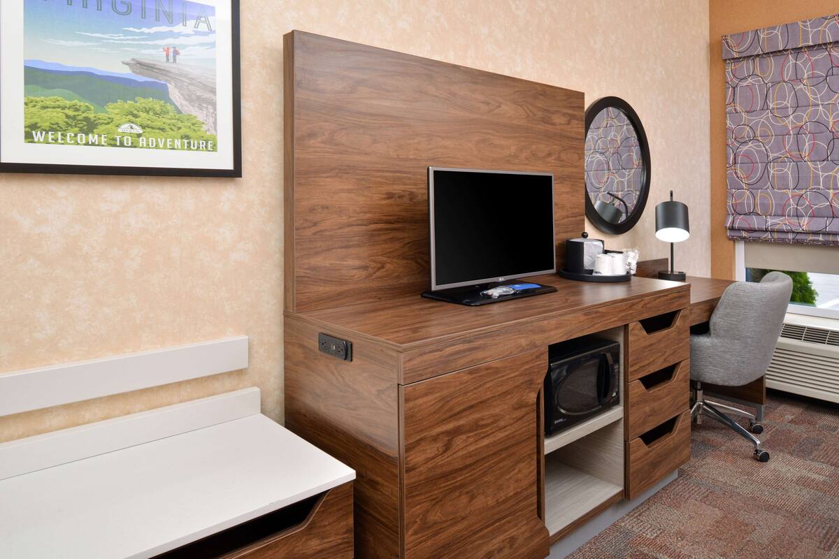 A hotel room with a wooden desk, a flat-screen TV, a microwave, a chair, and a large window with a view of the outdoors.