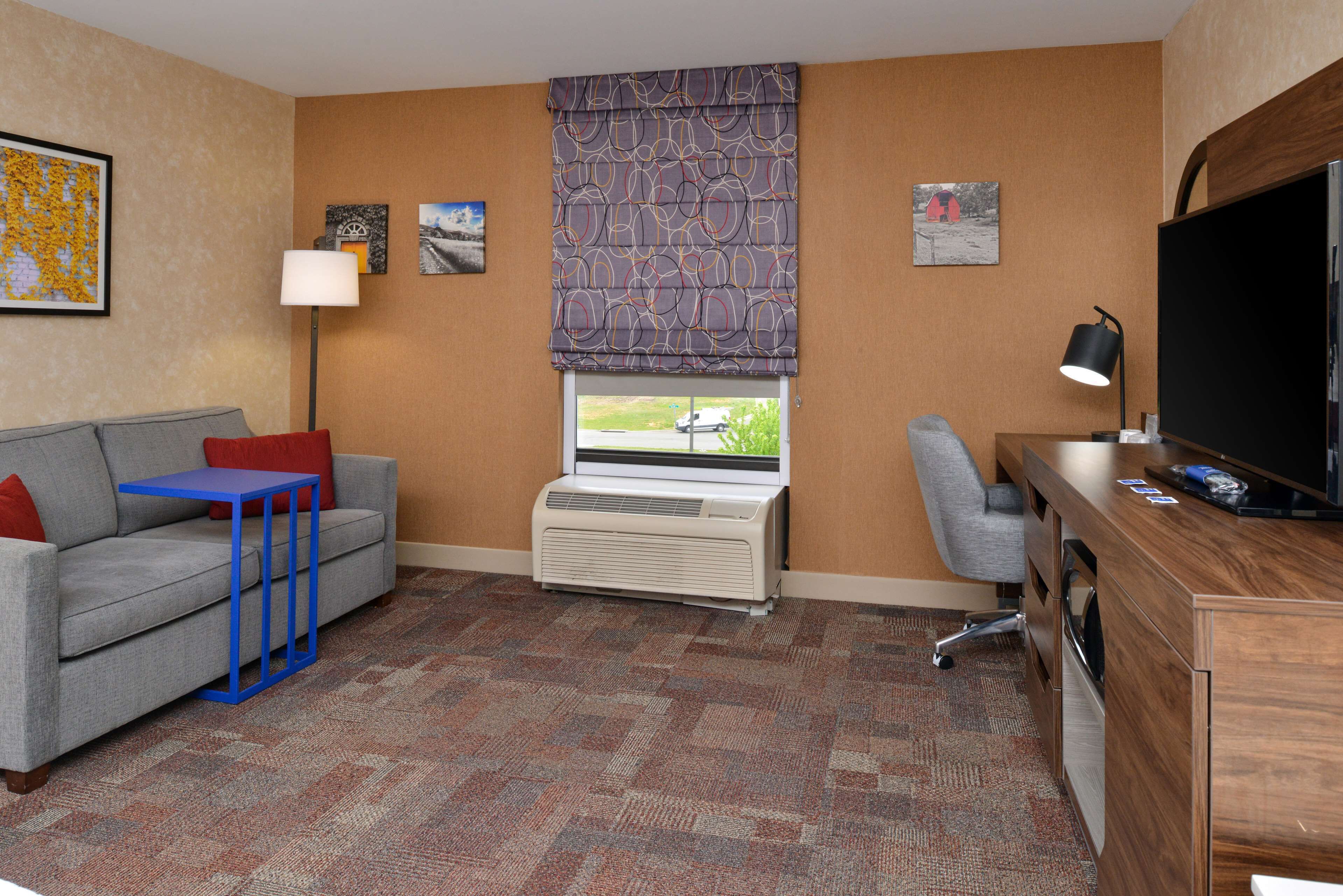 A hotel room with a gray sofa, a small blue table, a desk with a TV, a heater, and a window with a patterned blind.