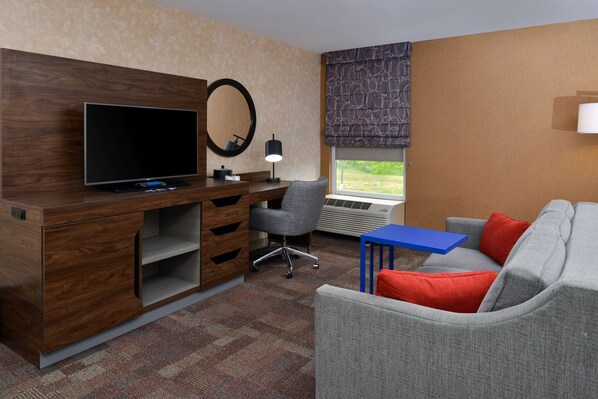 A hotel room with a wooden desk, a television, a grey office chair, a blue table, a grey sofa with red cushions, and a window with a view of greenery. - Hampton Inn Wytheville (Wytheville)