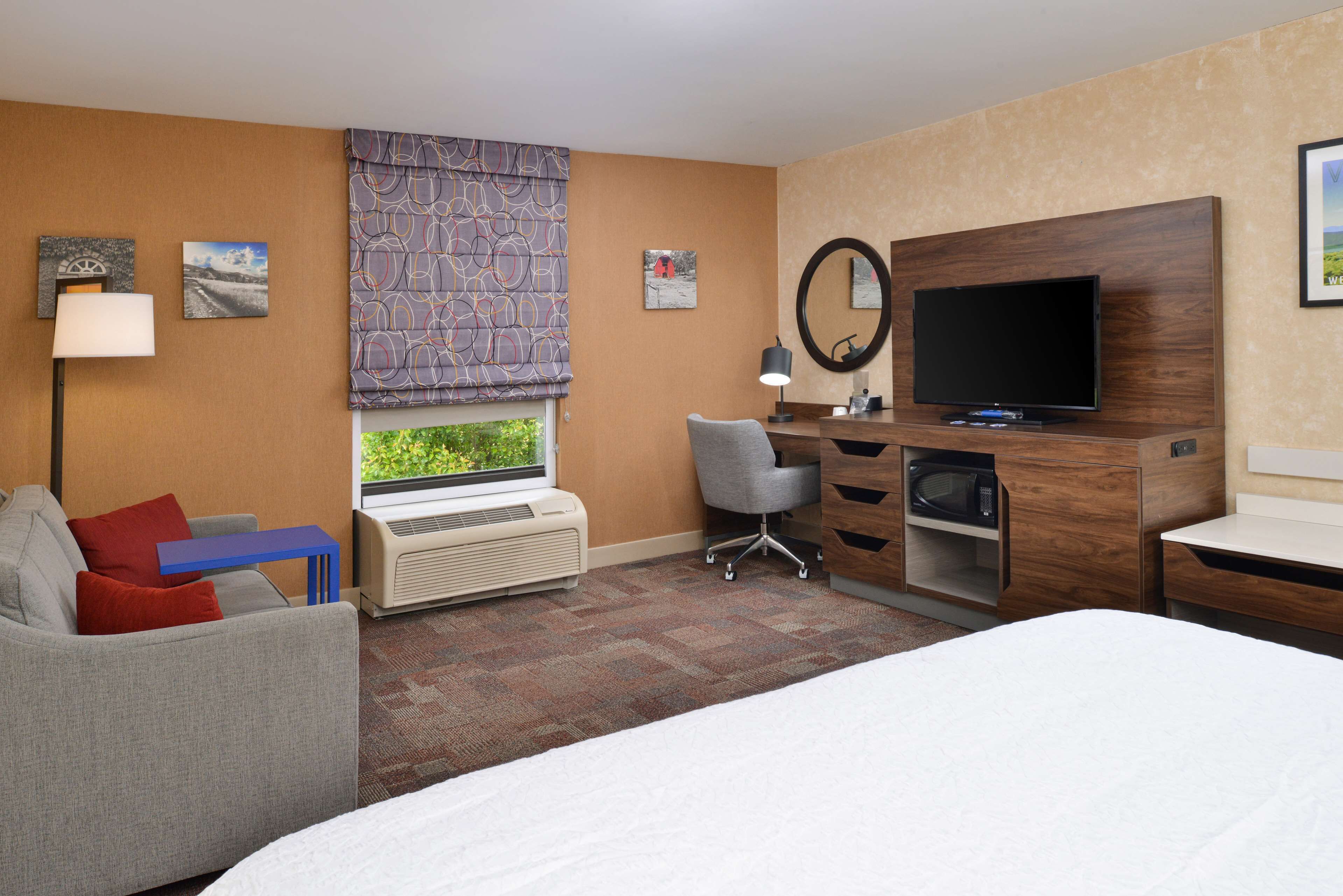 Une chambre d’hôtel comprenant un lit, un bureau avec un téléviseur à écran plat, un fauteuil de bureau gris, une table d’appoint bleue, un canapé gris avec des coussins rouges, une fenêtre donnant sur un espace verdoyant et un climatiseur fixé au mur.