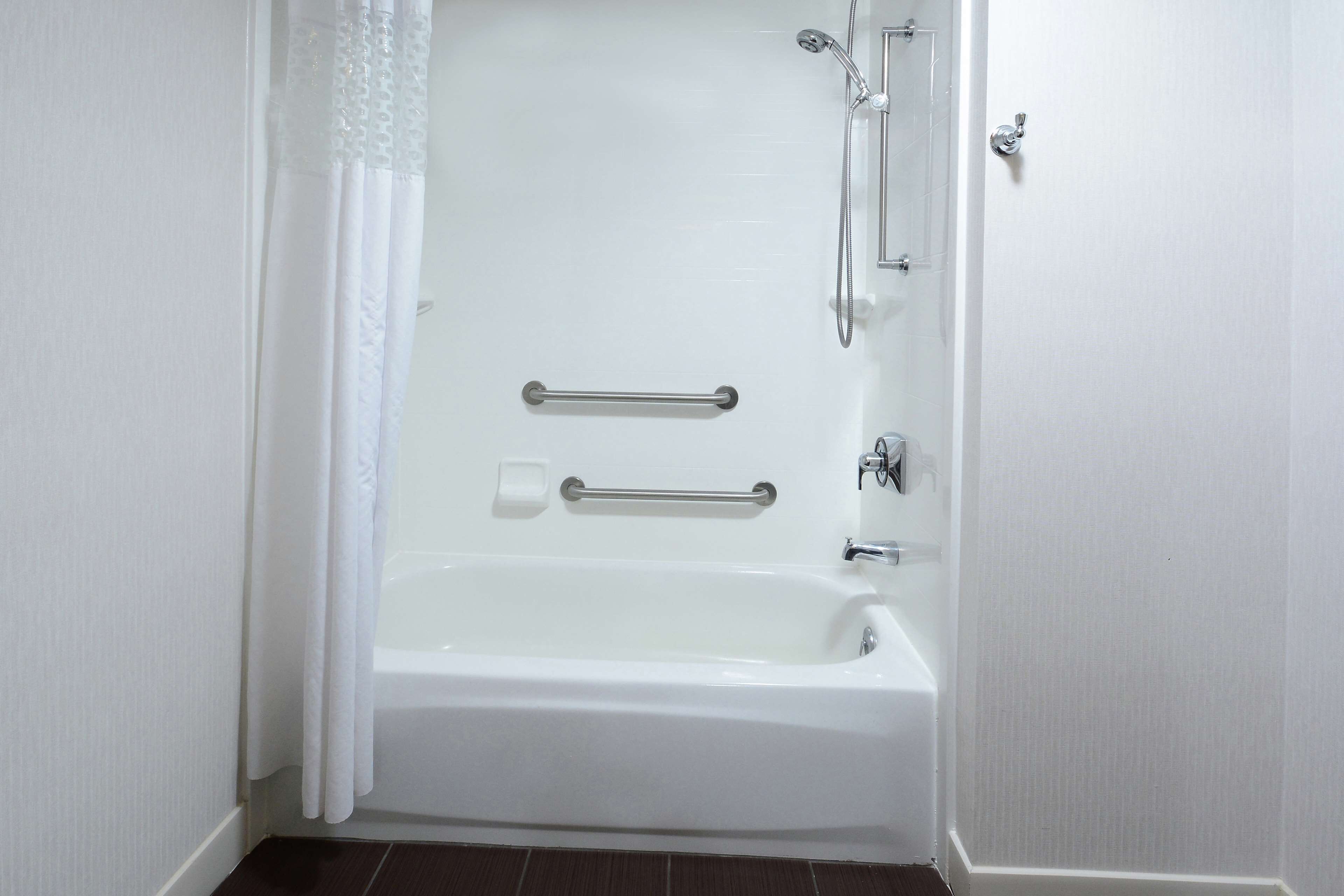 Una bañera blanca con ducha, barras de apoyo y un cabezal de ducha manual.
