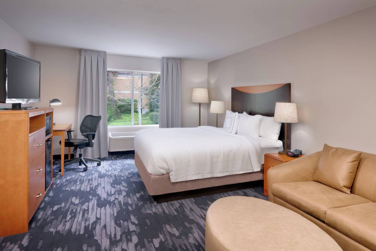 A hotel room with a bed, a desk with a TV, a chair, a sofa, and a window with curtains.