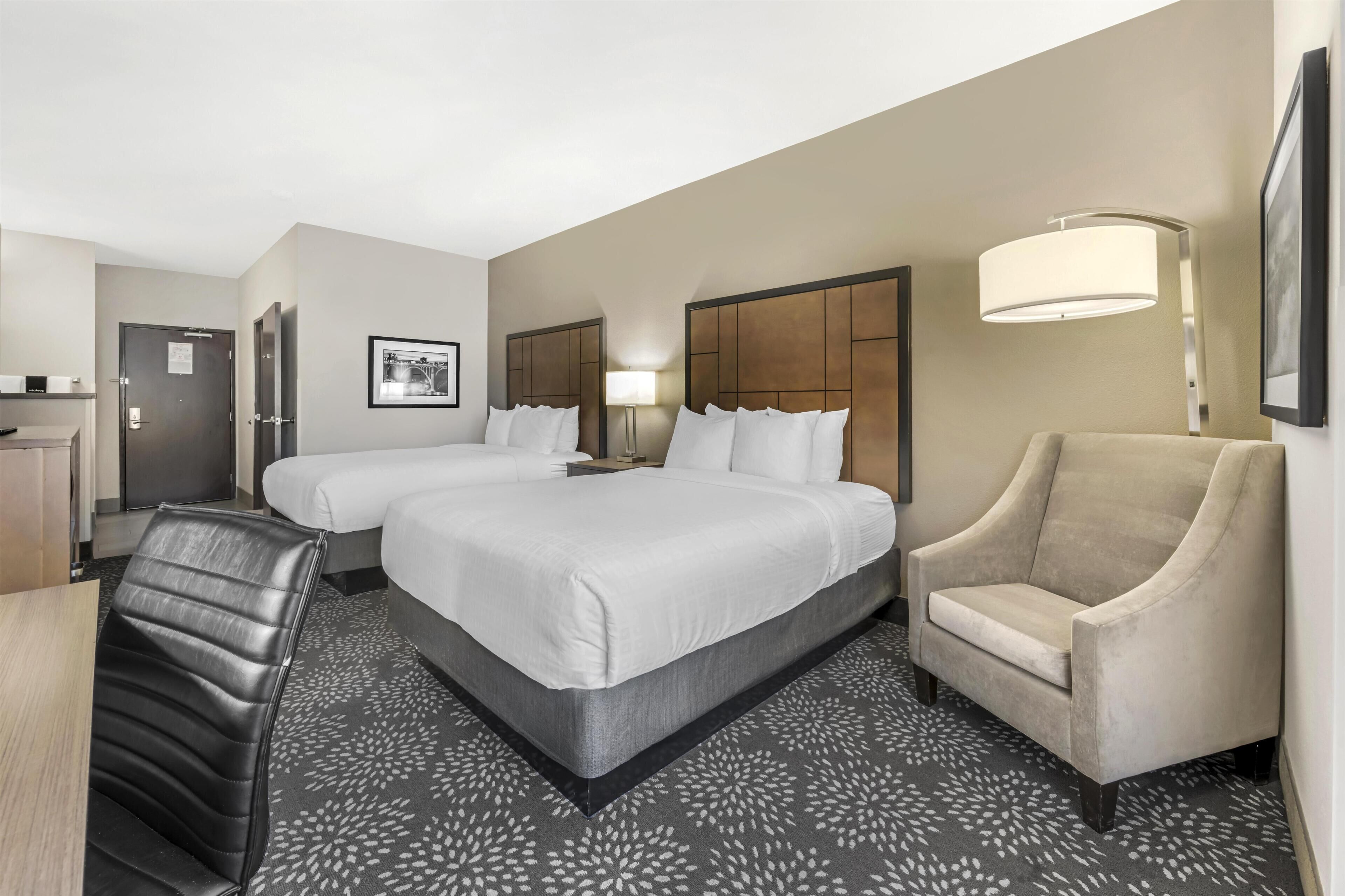 A hotel room with two beds, a desk, a chair, and a lamp.