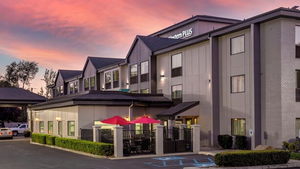A modern hotel with a sign reading "Western Plus," featuring a gated entrance, outdoor seating with red umbrellas, and a parking lot.