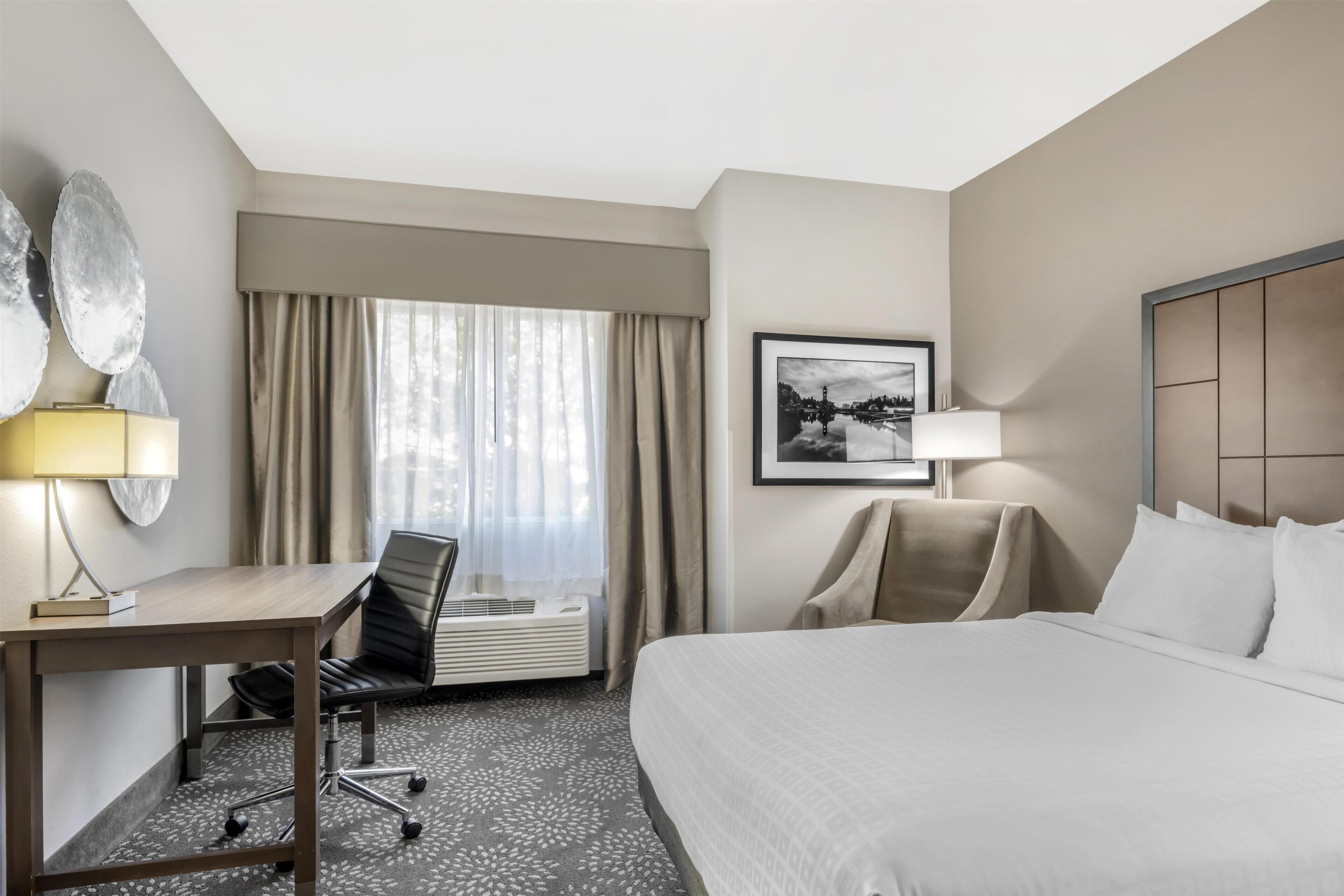 Una habitación de hotel con cama, escritorio, silla y un ventanal con cortinas.