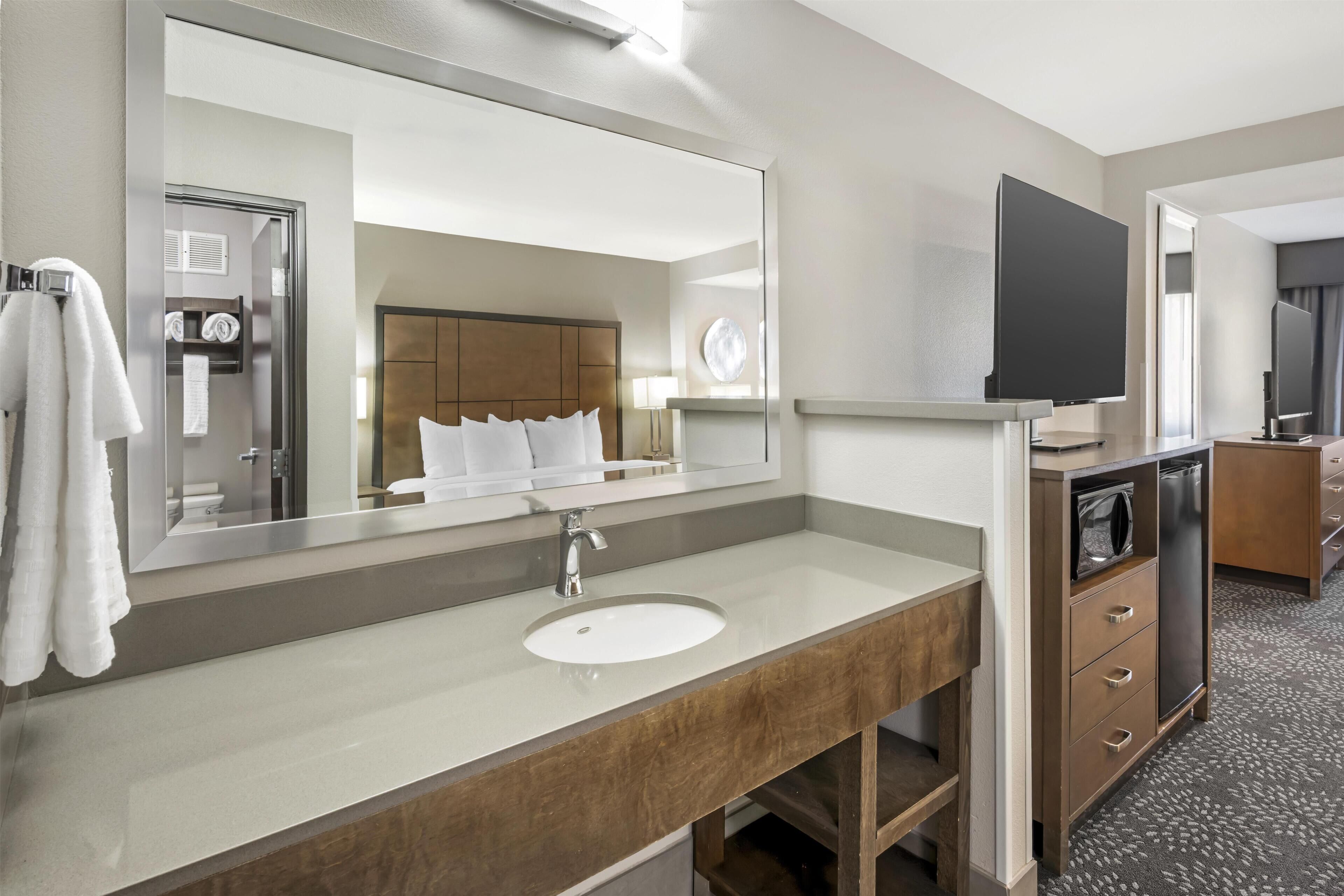 A hotel bathroom with a large mirror, a sink, a vanity, a television, and a shower area.