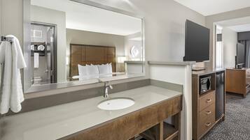 A hotel bathroom with a large mirror, a sink, a vanity, a television, and a shower area.
