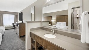 A hotel bathroom with a large mirror, a sink, a vanity, and a bed in the adjacent room.