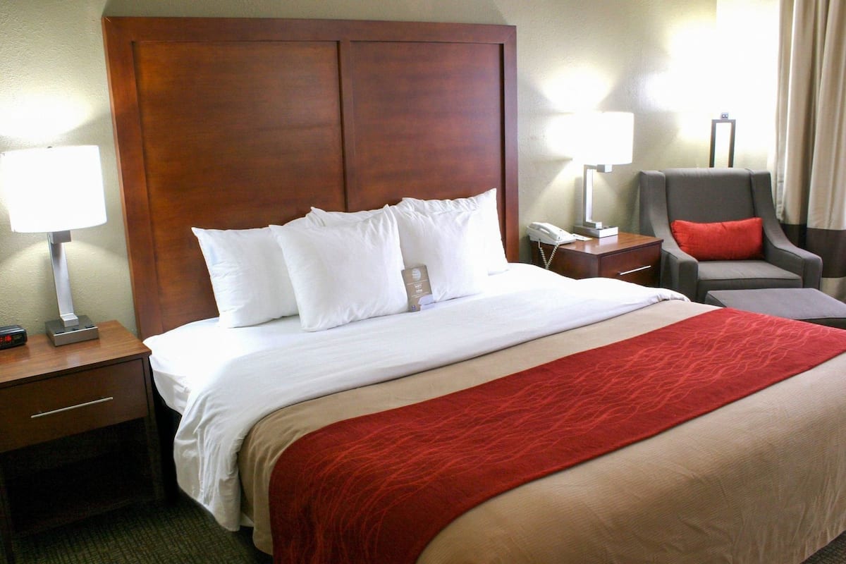 Una habitación de hotel con una cama grande, dos mesitas de noche, una silla y una lámpara.