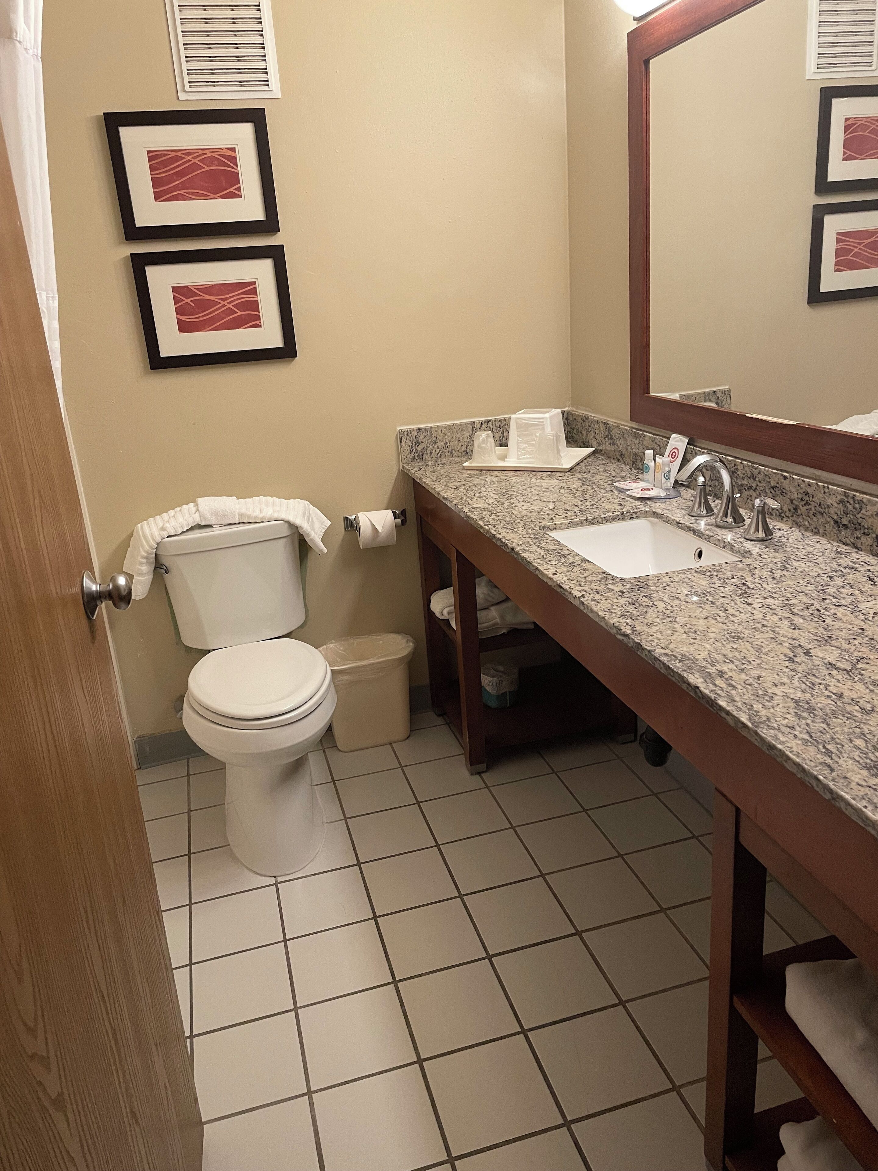 A bathroom with a toilet, sink, and mirror. There are framed pictures on the wall and a trash can.