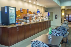 A breakfast bar with a refrigerator, coffee machine, and various food items. There are multiple seating areas with patterned chairs and tables.
