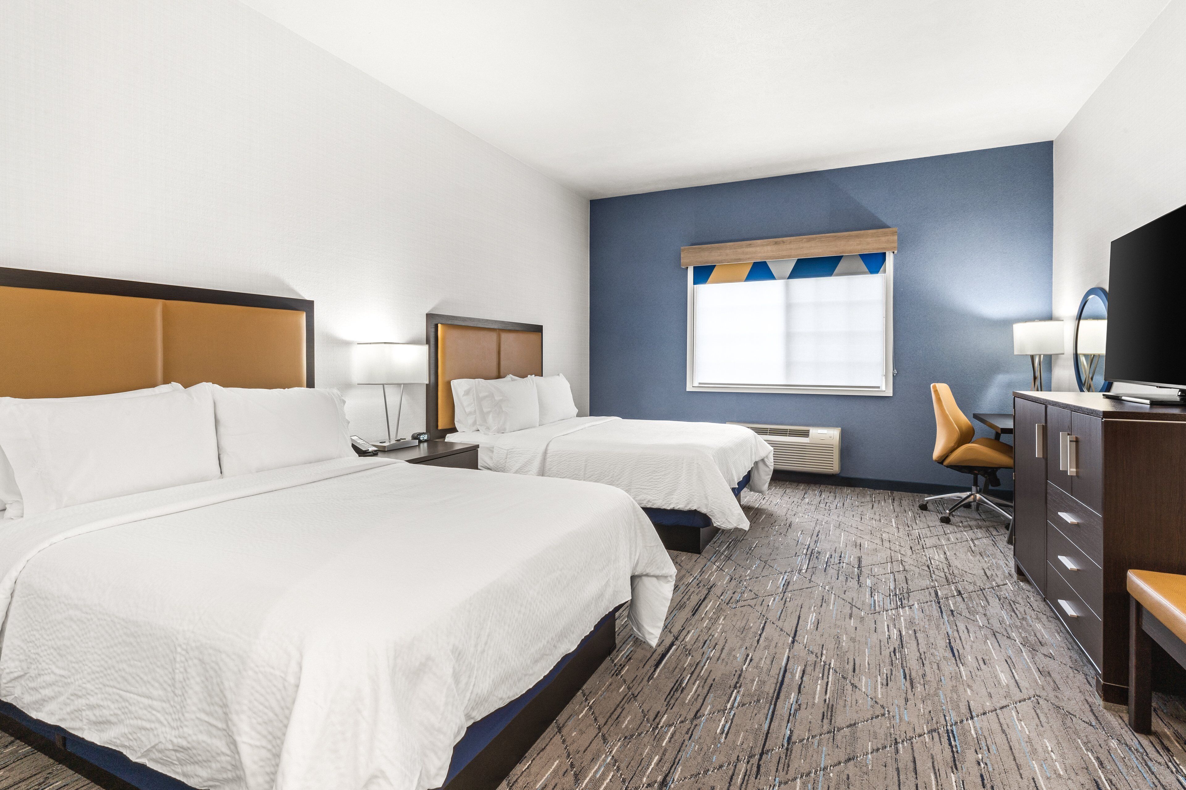 A hotel room with two beds, a desk, a chair, a dresser, a window with a view, and a lamp.
