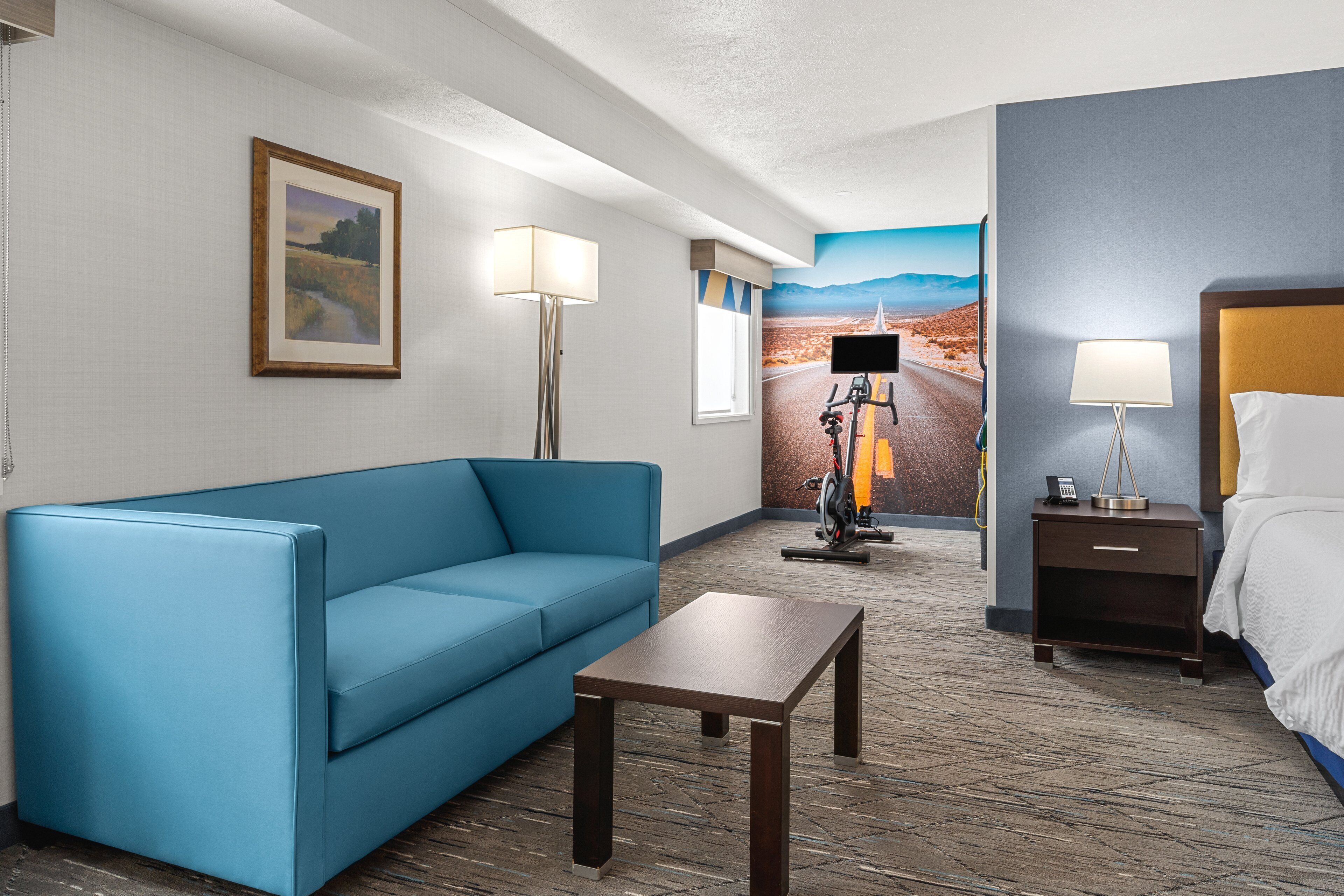 A hotel room with a blue sofa, a small wooden table, a bed with white linens, a nightstand with a lamp, and a wall-mounted TV.
