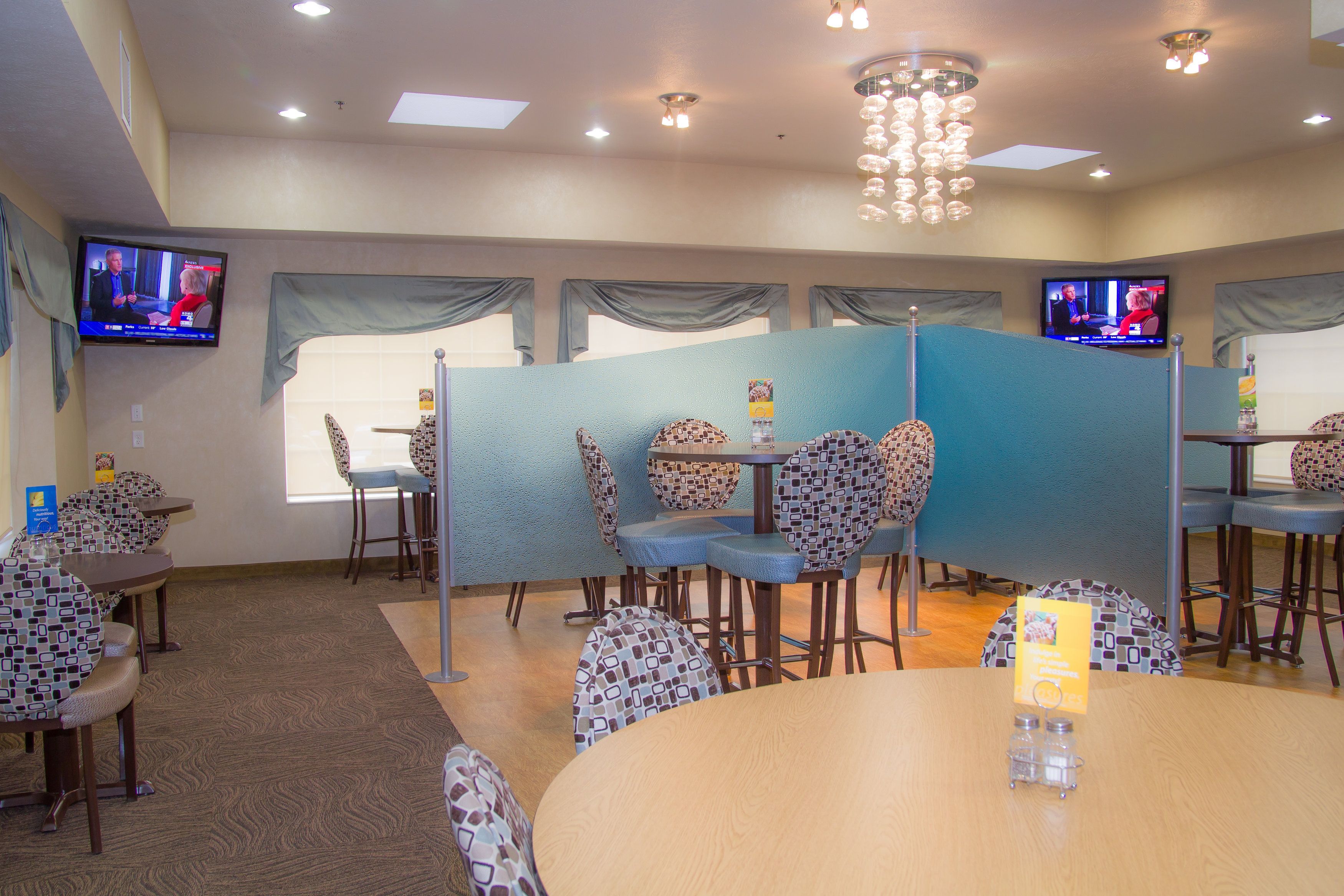 A conference room with round tables and patterned chairs, two televisions on the walls, and a chandelier.