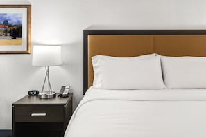A neatly made bed with white linens, a bedside lamp, a clock, and a phone on a wooden nightstand.