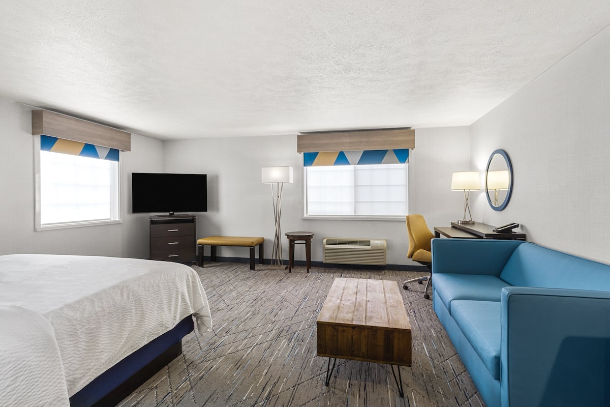 A hotel room with a bed, a blue sofa, a small table, a television, a lamp, and a mirror.
