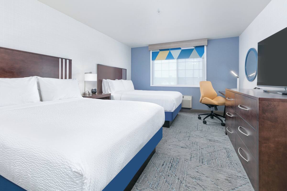 A hotel room with two beds, a desk, a chair, a TV, and a window with a geometric curtain.