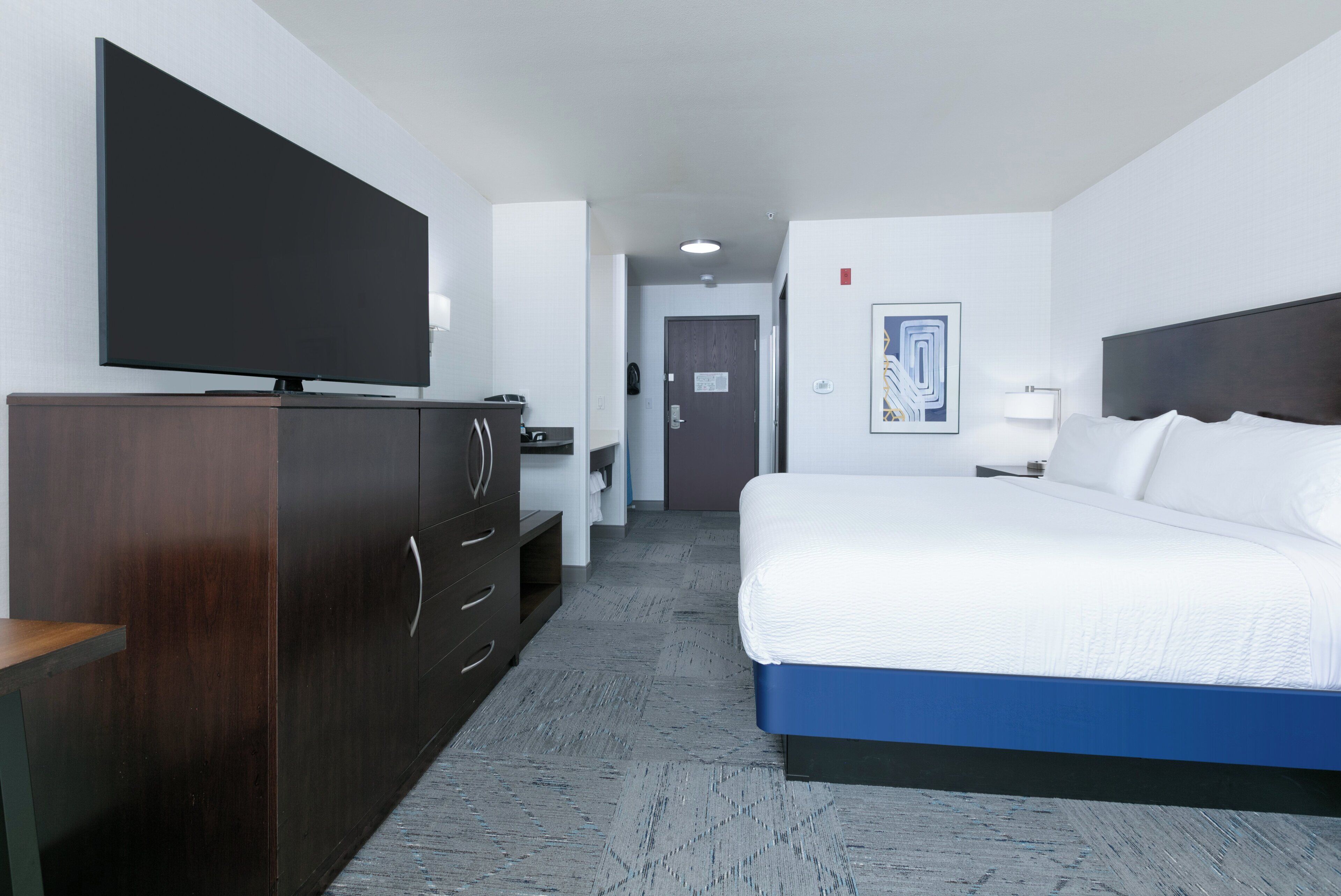 A hotel room with a large bed, a TV on a wooden dresser, a desk, and a door leading to another room.