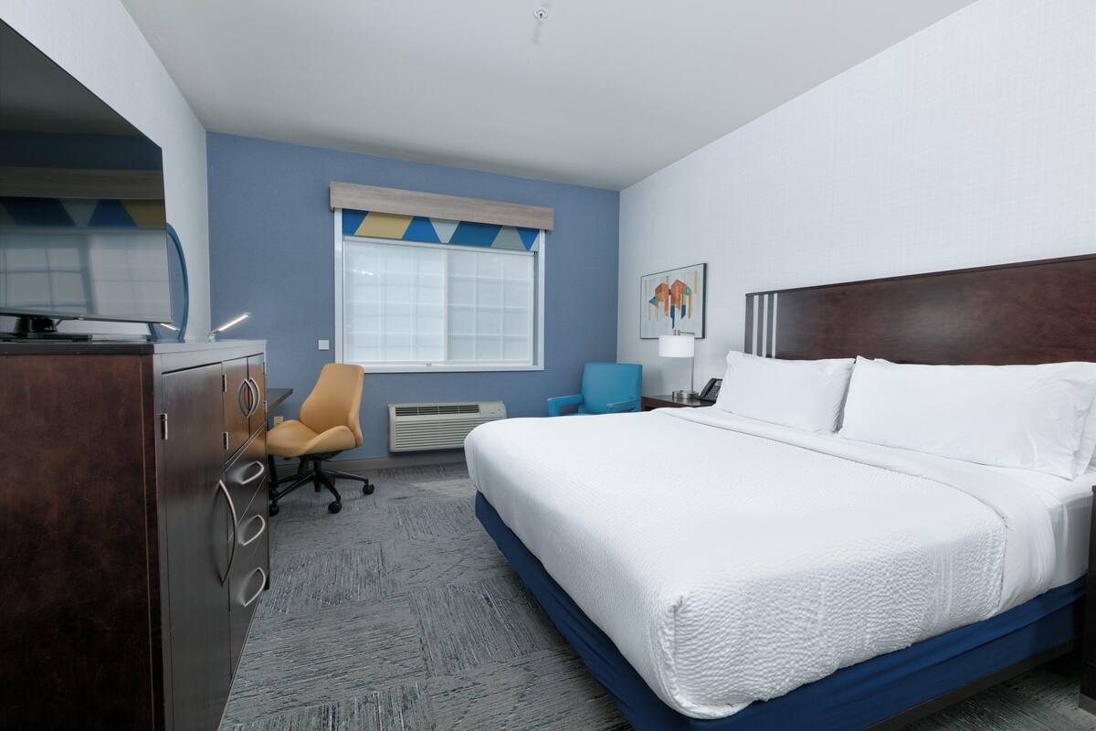 A hotel room with a large bed, a desk, a chair, and a window with blinds.