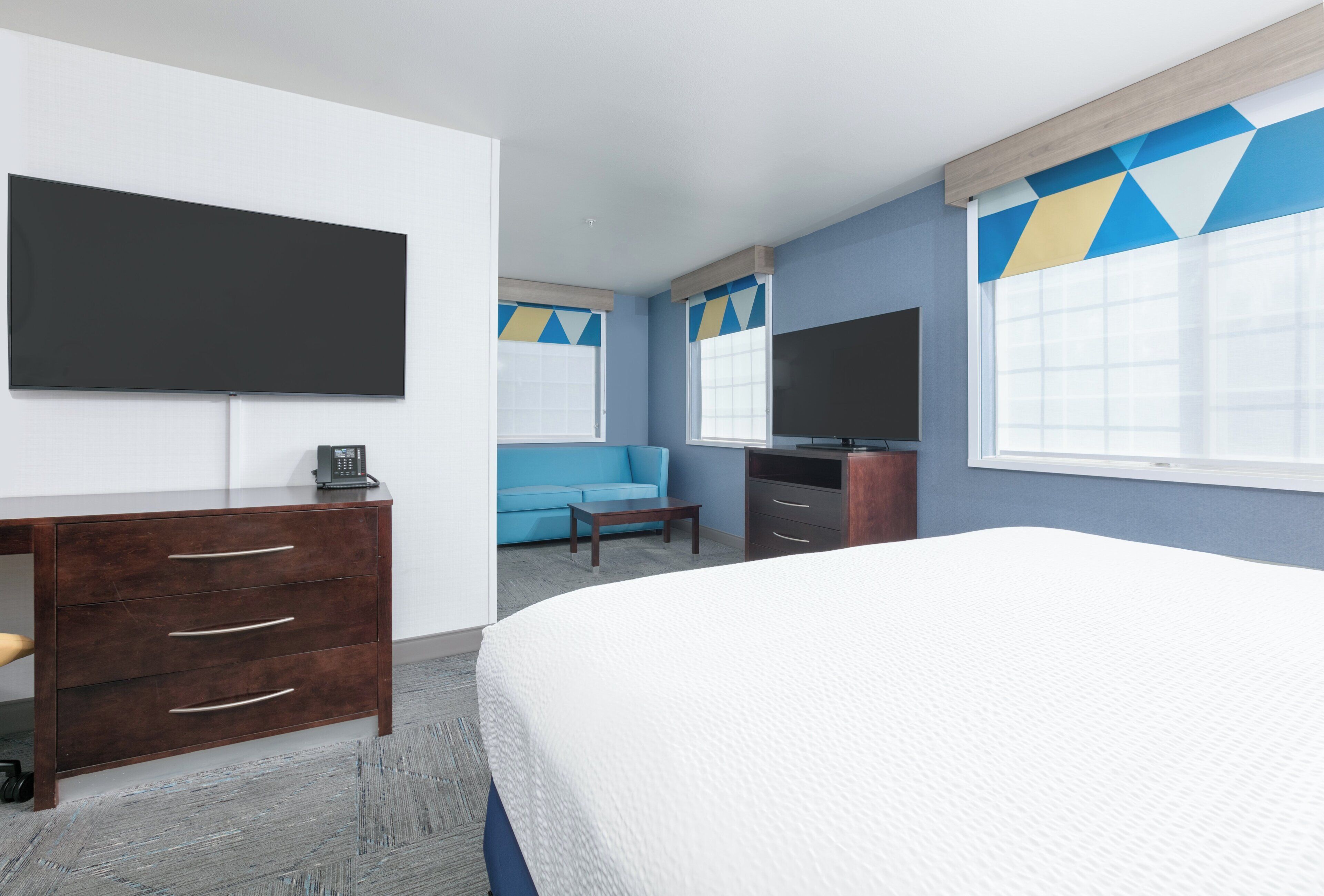 A hotel room with a large bed, a desk with drawers, a flat-screen TV, a blue sofa, and a window with geometric curtains.