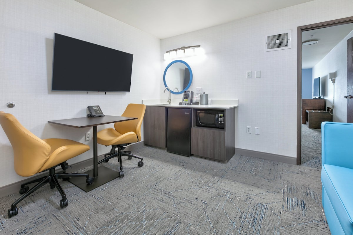 A modern hotel room with a flat-screen TV, a desk with two chairs, a microwave, a sink, and a blue couch.