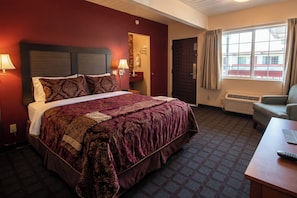 A hotel room with a large bed, a desk, a chair, a window, and an air conditioning unit.