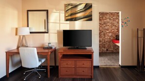 A hotel room with a wooden desk, a television, a white chair, a mirror, and a brick wall. - Travelers Inn by OYO Longview Kelso (Longview)