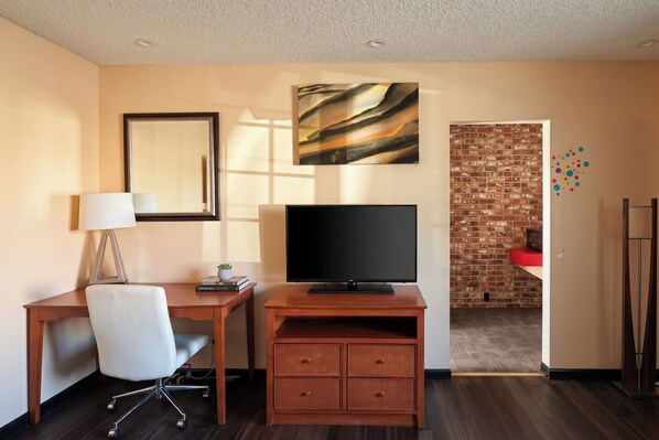 A hotel room with a wooden desk, a television, a white chair, a mirror, and a brick wall.