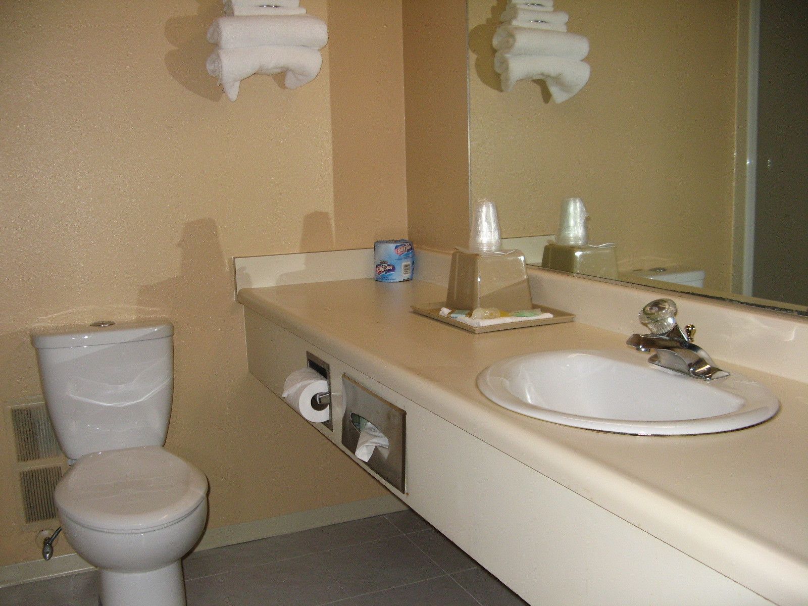 Un baño con inodoro, lavabo y espejo. Hay toallas colgadas en la pared, un dispensador de papel higiénico y un dispensador de jabón sobre el lavabo.