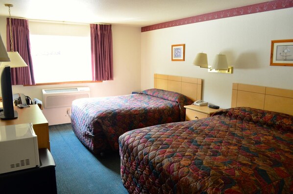 A hotel room with two beds, a desk with a lamp, an air conditioner, and a window with curtains.