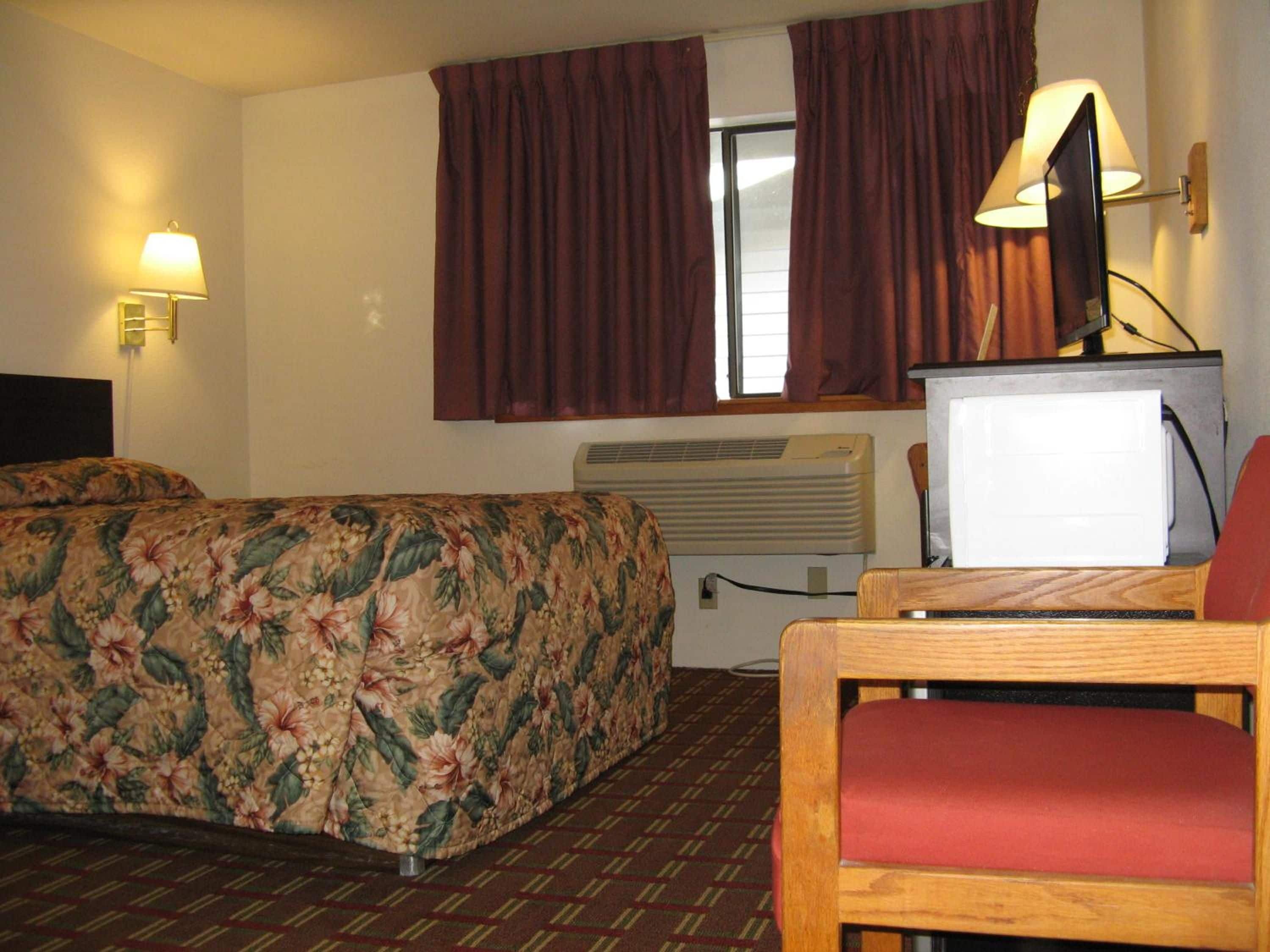 Ein Hotelzimmer mit Bett, Schreibtisch mit Computer, Stuhl, Fenster mit Vorhängen und einer Wandlampe.