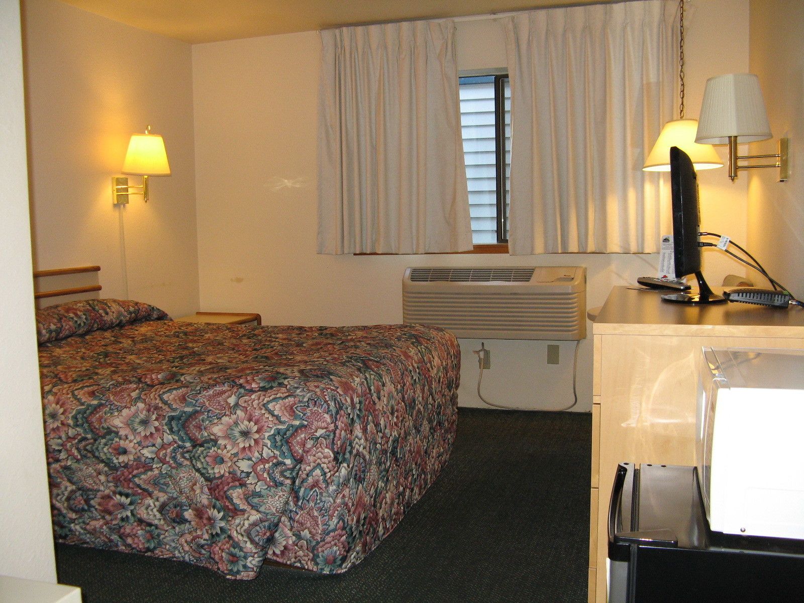 A hotel room with a bed, two wall-mounted lamps, an air conditioner, a desk with a lamp, and a window with curtains.