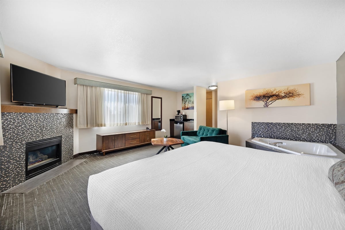 A hotel room with a large bed, a flat-screen TV mounted on the wall, a fireplace, a desk, a green armchair, a sofa, and a wall-mounted lamp.