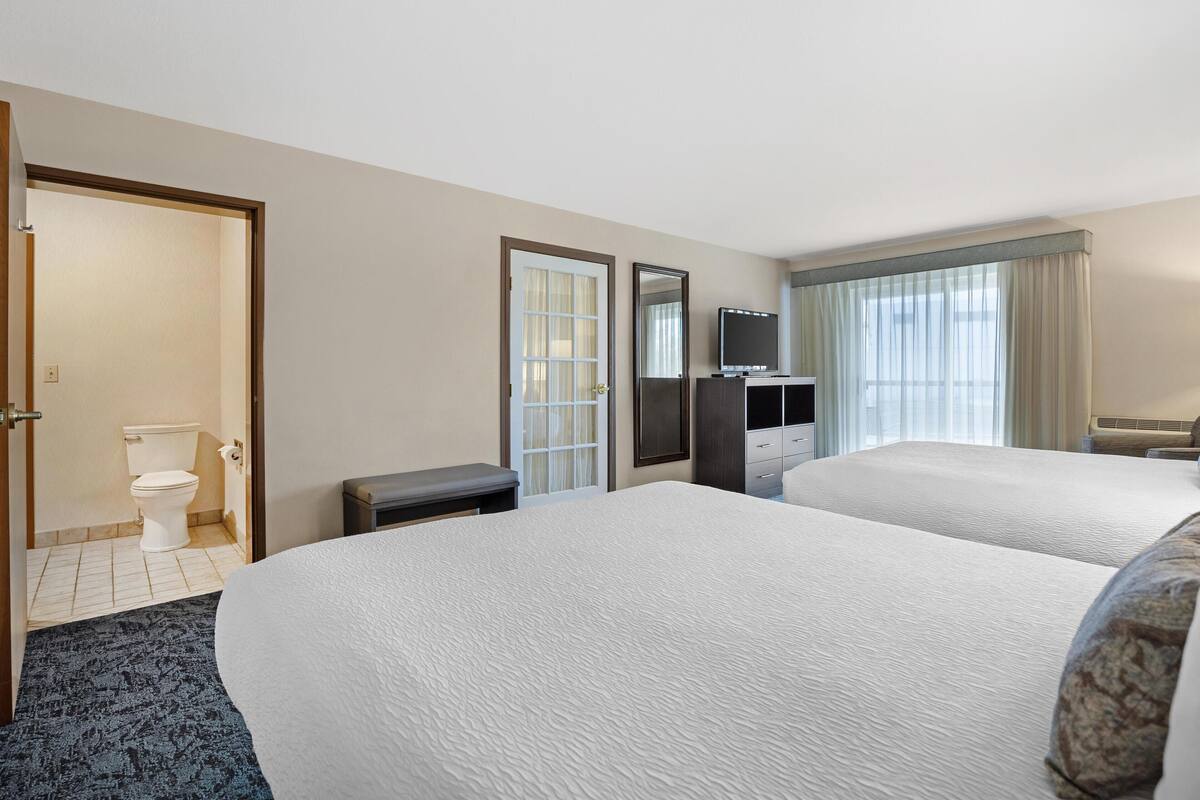 A hotel room with two beds, a TV, a desk, and a bathroom visible through an open door.