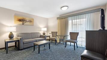 Habitación de hotel con sofá, dos sillones, una mesa de centro, lámpara, televisor y un ventanal con cortinas.