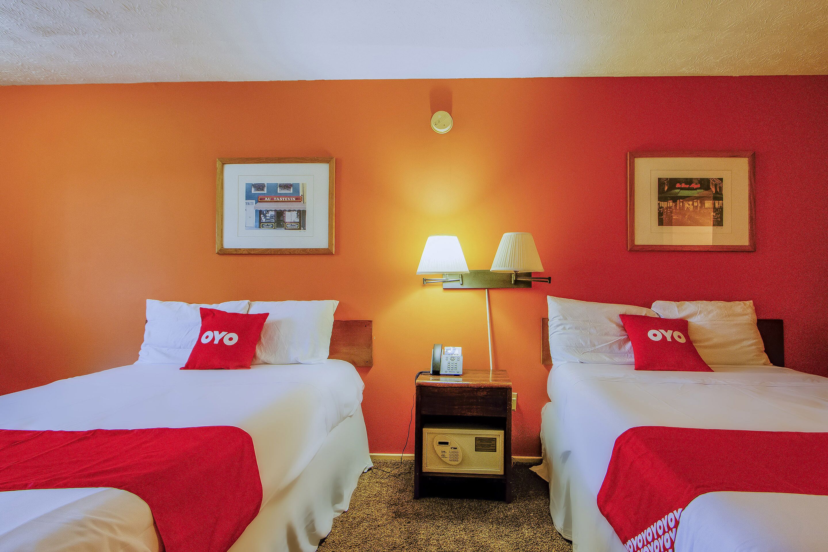 Quarto de hotel com duas camas, cada uma com almofada vermelha da OYO, roupa de cama branca e mesa de cabeceira com abajur e telefone.