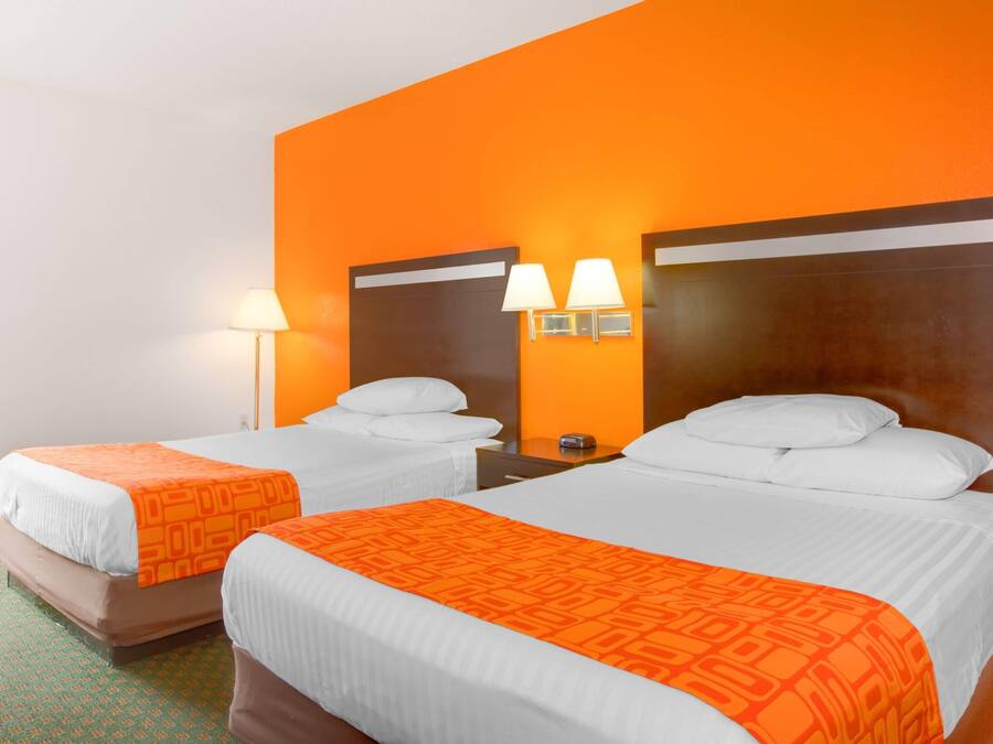 Et hotelværelse med to senge, begge med hvide sengetæpper og orange pyntepuder, et natbord med lampe, samt en vægmonteret lampe.