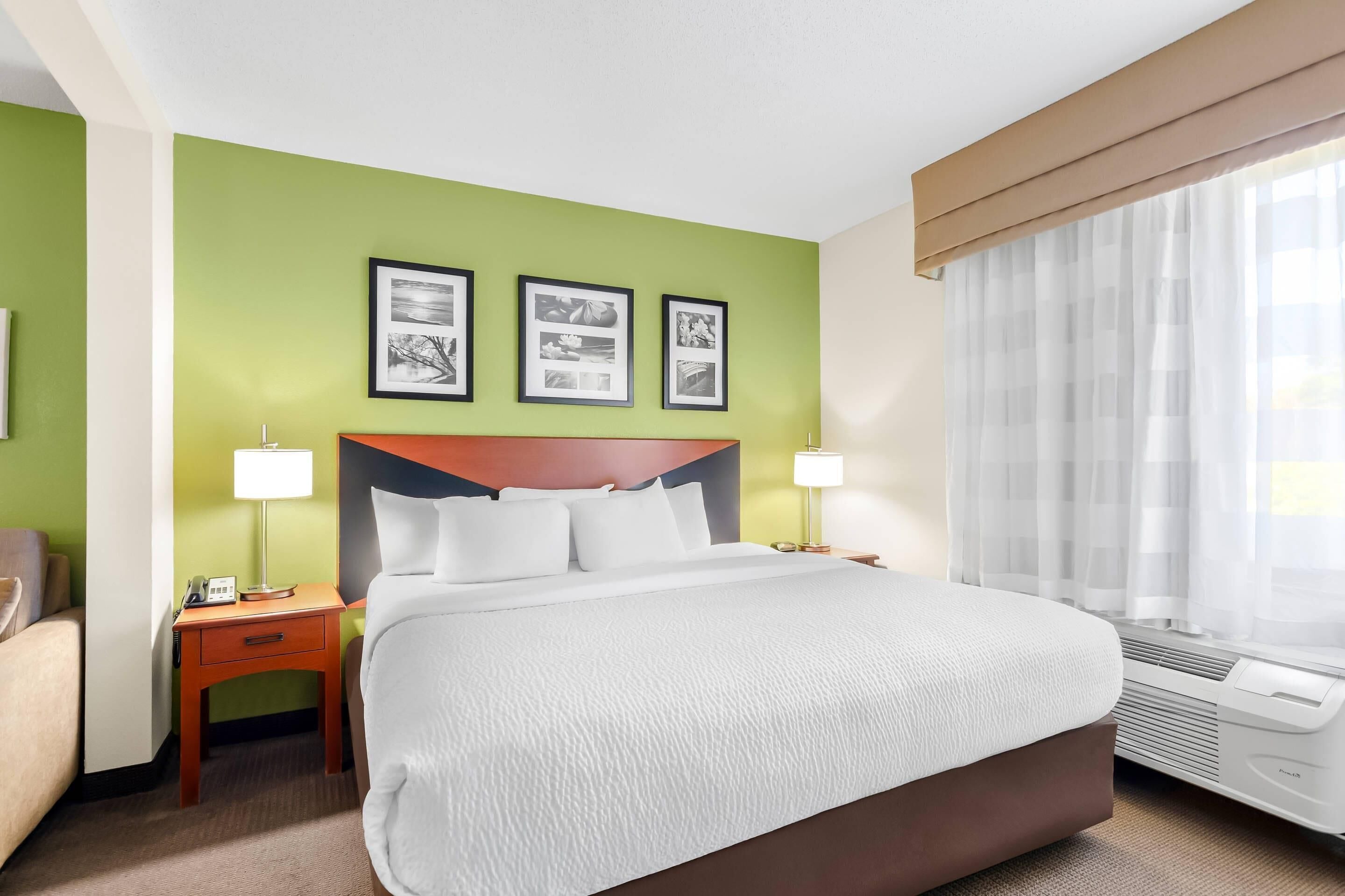 A hotel room with a large bed, two bedside tables with lamps, a wall-mounted air conditioner, and three framed pictures on the wall.