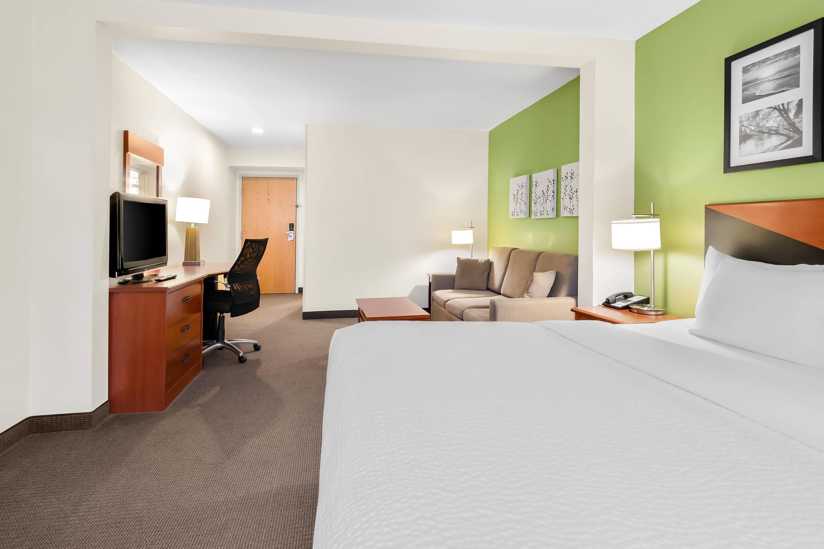 A hotel room with a large bed, a desk with a computer, a chair, a sofa, and a TV.