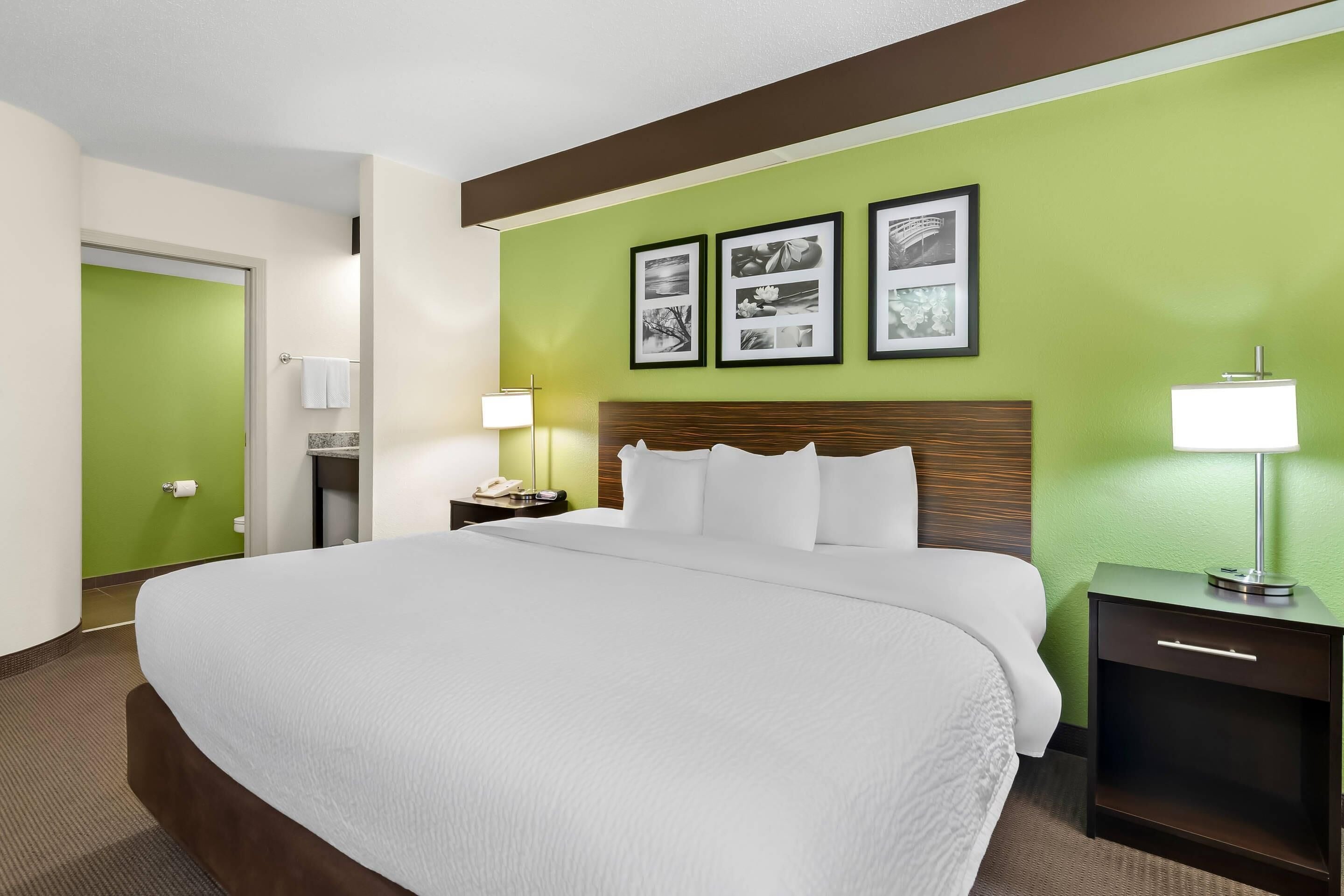 Habitación de hotel con una cama grande, una mesita de noche con lámpara, una pared de acento verde y cuadros enmarcados sobre la cama.