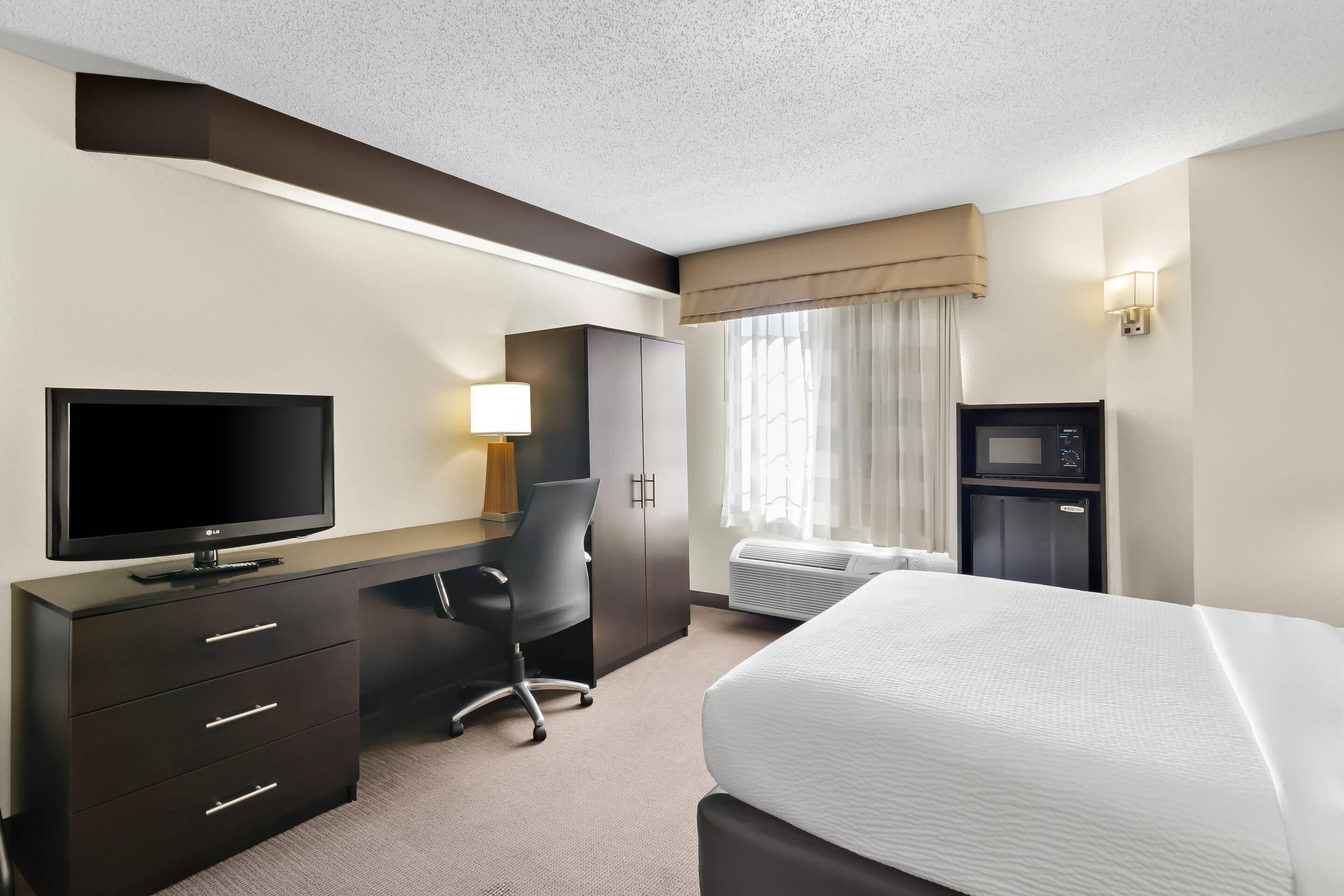 Habitación de hotel con cama, escritorio con televisor, silla, lámpara, microondas y ventana con cortinas.
