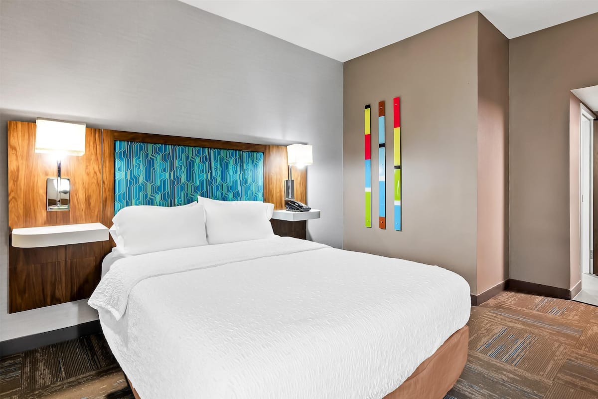 Ein Hotelzimmer mit einem großen Bett, einem Holz-Kopfteil, einem Nachttisch mit Lampe und einem farbenfrohen, abstrakten Kunstwerk an der Wand.